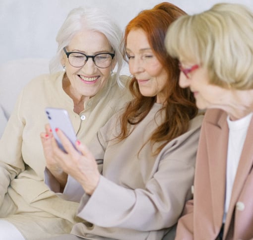 Three older women together, the lady in the middle is holding a phone video chatting while the other two  ladies each side of her are joining in