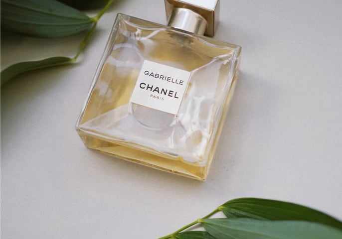 Gabrielle perfume bottle laid down on it's side, logo facing up with green leaves above and below it.